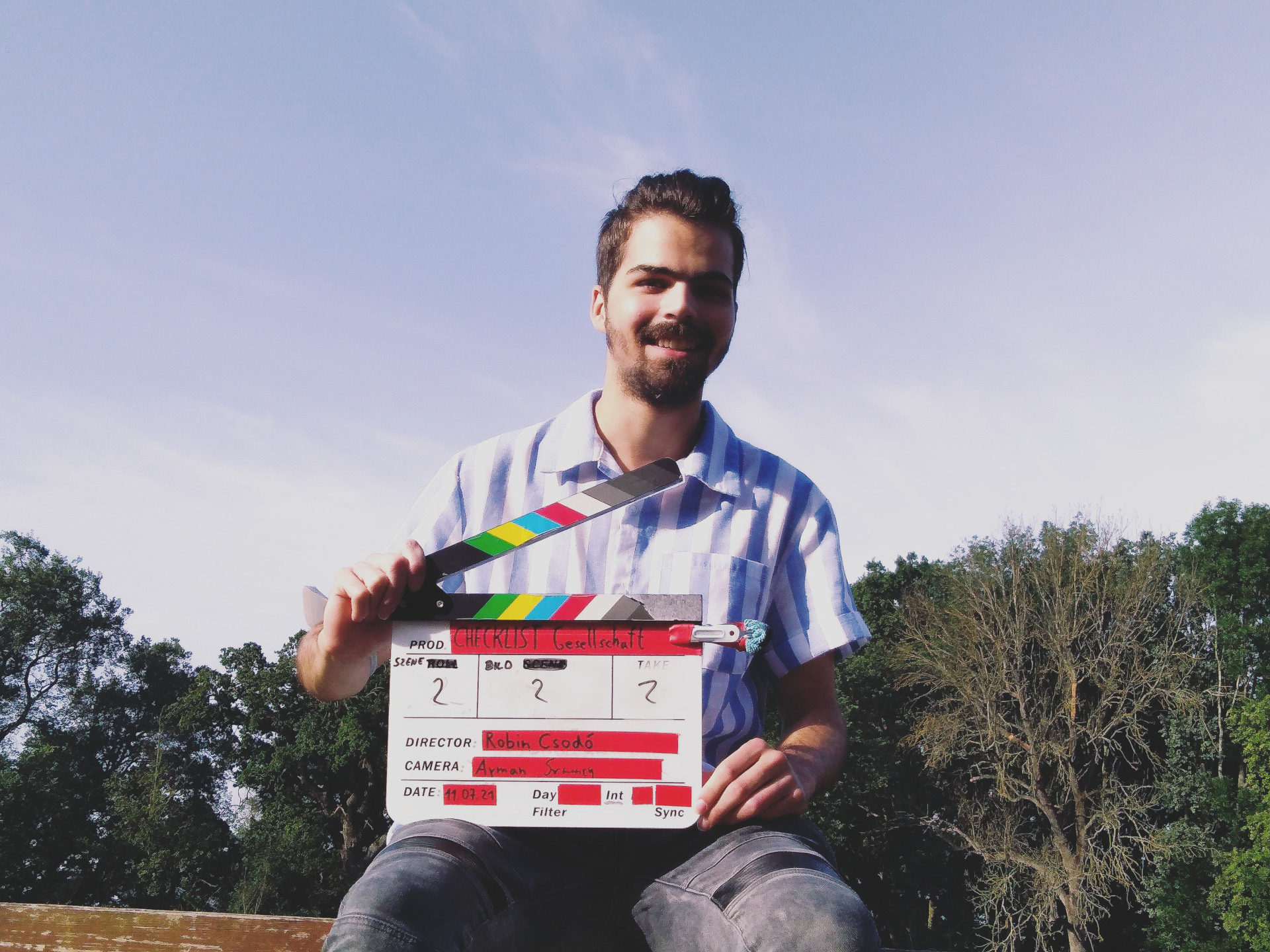 ESC Projektélmények Németországban A képen egy fiatalember filmes csapótáblával a kezében ül egy kerítés tetején