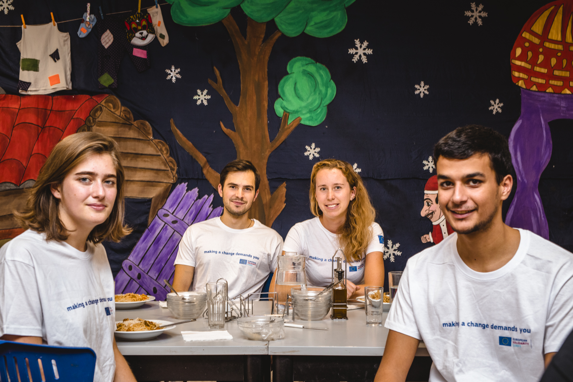 Rajzokkal kifestett fal előtt egy asztalnál ül 4 fiatal és néz a kamerába making a change demands you feliratú pólóban