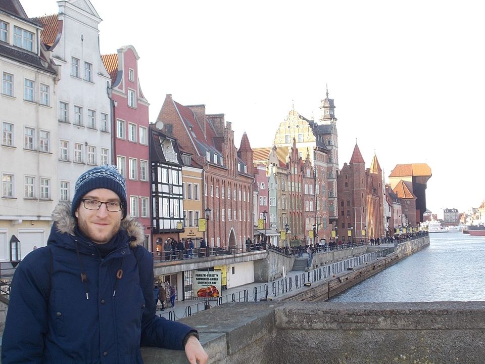 Őszi hangulatú képen áll Gósi János Gergő Gdansk folyój fölött húzódó kőhíd pillérénél a háttérben különleges házak sora