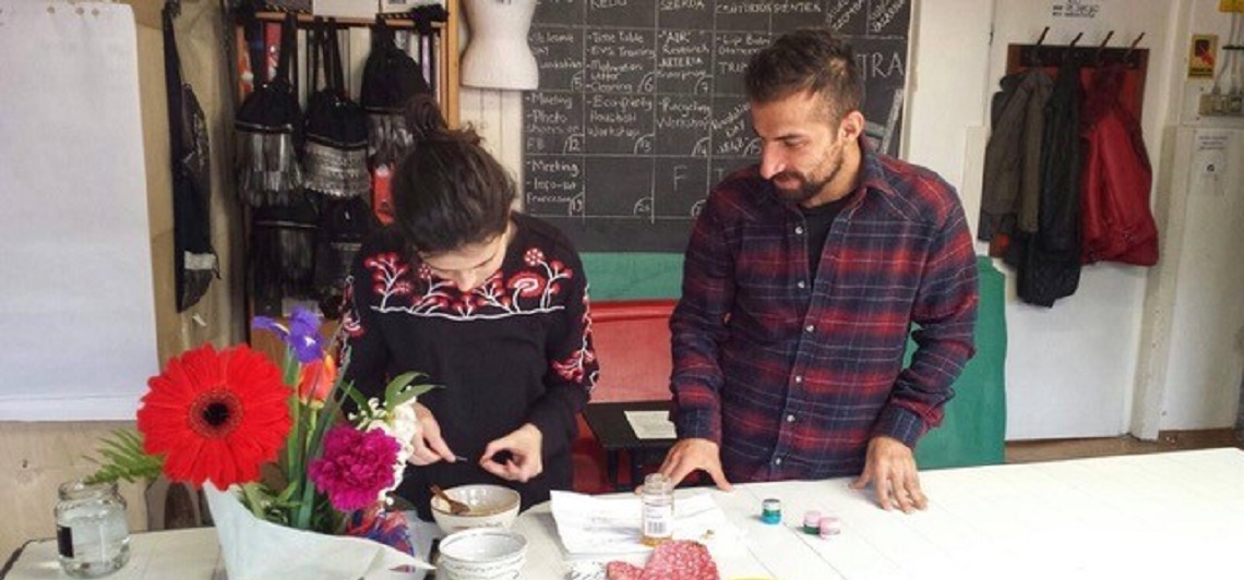 Egy hosszú asztal mellett kézműveskedik egy férfi és egy nő körülöttük virágok mögöttük a falon tábla és kabátok a falon